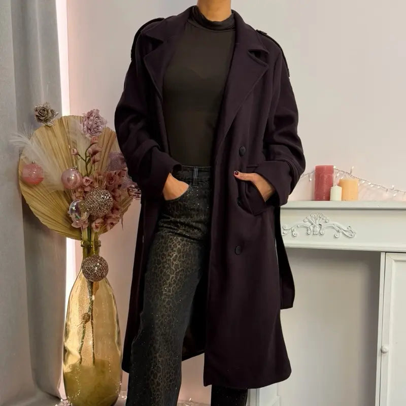 Ce manteau croisé présente une fermeture boutonnée, une ceinture intégrée et des poches pratiques, alliant élégance raffinée et praticité polyvalente.
